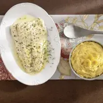 Fotografia em tons de amarelo em uma bancada de madeira com uma toalha florida, um prato oval branco com o peixe temperado com tomilho. Ao lado, um potinho branco com o purê de banana.
