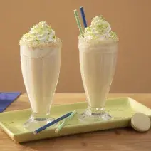 Foto da receita de Milkshake de Bono Torta de Limão Coberto, em dois copos decorados com chantilly, sobre um prato verde, em uma bancada de madeira decora com fatias de limão, biscoitos brancos e um tecido em tom azul