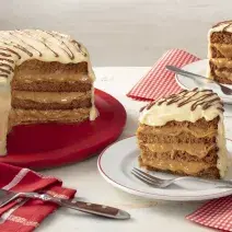 Fotografia em tons de vermelho em uma bancada cinza com paninhos vermelhos listrados e de bolinhas brancas. Ao centro, um prato branco com uma fatia do bolo de pão de mel com chocolate branco e à esquerda, um suporte grande vermelho com o bolo inteiro.
