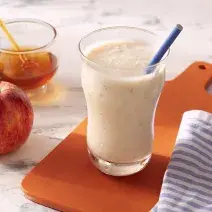Fotografia em tons de branco em uma bancada de madeira de cor branca. Ao centro, uma tábua de madeira laranja com um copo em cima contendo a bebida. Ao lado, há um pano branco listrado, 1 maçã e um recipiente com mel.