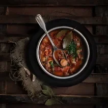 Fotografia vista de cima de uma sopa com linguiça, feijão, cenoura, costela e duas folhas de loura por cima. Apoiada na sopa, uma colher de sopa, que está dentro de um recipiente fundo de alumínio, sobre um prato preto, que está sobre uma mesa de madeira.