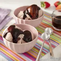 Foto da receita de Sorvete de Morango Caseiro com Calda de Leite Moça e Nescau. Observa-se dois recipientes com duas bolas de sorvete e a calda de chocolate por cima. Morangos decoram a foto.