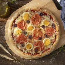 Fotografia em tons de azul em uma bancada de madeira escura, um paninho azul, uma tábua de madeira com a pizza portuguesa em cima dela. Ao lado, um potinho de azeite e um rolo de massa.