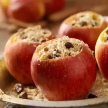 Fotografia de maçãs assadas com uva passa, aveia e mel dentro de uma forma redonda de alumínio, que está sobre um pano branco. Ao fundo, algumas maçãs inteiras sobre uma tábua de madeira.
