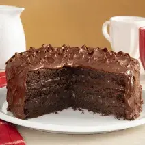 Fotografia em tons de vermelho em uma bancada de madeira com um pano vermelho listrado em branco, com um prato branco no centro com o bolo brownie com brigadeiro de cacau de cobertura. Ao fundo, uma xícara branca, uma xícara vermelha e um bule branco.