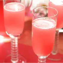Fotografia em tons de rosa em uma bancada de madeira, um pano vermelho, taças de vidro de champagne com as bebidas feitas com Picolé de Limão com espumante.
