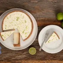 Fotografia em tons de branco e marrom de uma bancada de madeira vista de cima, com um prato branco com uma torta de limão fatiada, ao lado direito prato com uma porção de torta com um garfo e limões, e no esquerdo um guardanapo azul.