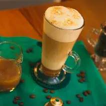 Fotografia em tons de verde de uma mesa de madeira com um paninho verde, uma xícara com a bebida e grãos de café decorando.