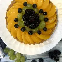 Foto vista de cima da receita de Torta de Frutas com Leite Moça, decorada com pêssegos, uvas, blueberrie, sobre um prato branco em uma bancada de madeira.