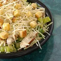 fotografia em tons de azul, preto, amarelo e verde de uma bancada azul vista de cima. Contém um recipiente redondo preto com a salada, e pedaços de frango e queijo ralado.