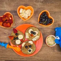 Fotografia em tons de laranja em uma bancada de madeira escura, um prato laranja com três panquequinhas com rostinho de bichinhos em cada uma delas, potinhos com frutas e um ursinho azul de pelúcia.
