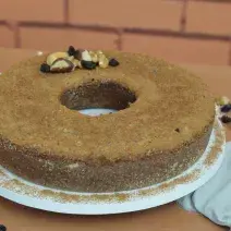 Fotografia em tons de marrom de uma bancada de madeira com um prato branco redondo, sobre ele um bolo de nozes. Ao lado um paninho branco com garfo e nozes.