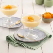 Fotografia em tons de verde em uma bancada de madeira branca, paninho verde, duas taças de vidro com a sobremesa de damasco e laranja dentro dela, enfeitadas com damasco. Ao lado, um potinho com alguns damascos.