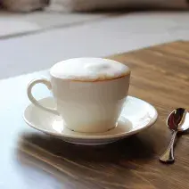 fotografia em tons de marrom e branco tirada de uma bancada marrom. Contém um pratinho branco com uma xicara de café branca, e dentro contém a bebida Cappuccino.