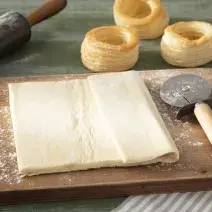 Fotografia em tons de marrom em uma bancada de madeira verde, uma tábua de madeira suja com farinha e um pedaço de massa folhada e um cortador de pizza ao lado.