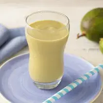 Fotografia em tons de azul e verde de uma bancada com um pratinho azul, um copo transparente com uma vitamina de abacate e um canudo. Ao fundo um paninho azul e um abacate.