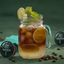 Fotografia em tons de verde de uma bancada verde, sobre ela um copo de vidro com cold brew e limões. Ao lado capsulas de Dolce Gusto Cold Brew e um paninho azul.