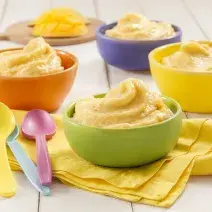 Fotografia em tons de amarelo em uma bancada de madeira de cor branca. Ao centro, um pano amarelo contendo uma tigela com o sorvete e ao lado, há algumas colheres coloridas. Ao fundo, mais alguns potes de sorvete.