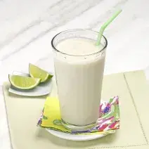 Fotografia em tons de verde em uma bancada de madeira cinza clara com um pano bege com detalhes verdes, um pratinho branco pequeno com um copo de vidro longo e grande com o smoothie de pera com limão. Ao lado, duas fatias de limão.