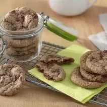Foto da receita de Cookies Sem Glúten. Observa-se um pote hermético de vidro com os cookies dentro sobre uma grade. Ao lado direito, cookies dispostos sobre um guardanapo verde.