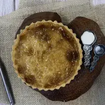 Foto vista de cima da receita de Torta de Leite Moça, sobre uma tábua de madeira, com duas colheres de ferro ao lado, uma delas com açúcar. Abaixo da tábua há um tecido e sobre ele uma faca do lado esquerdo.