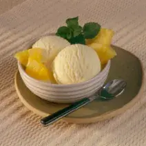 Fotografia com tons de bege e amarelo, ao centro bowl com duas bolas de sorvete de abacaxi, abacaxi fatiado, hortelão, sobre prato verde, ao lado uma colher de sobremesa, sobre toalha bege.