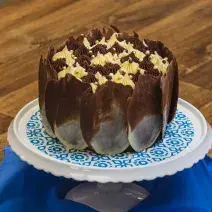 Foto aproximada de um bolo de chocolate com decoração de lascas marmorizadas, sobre um prato branco e azul em uma bancada de madeira