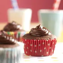 Fotografia em tons de branco e vermelho de uma bancada branca com bolinhas coloridas, sobre ela três cupcakes de Nescau. Ao fundo dois copos com canudos coloridos.