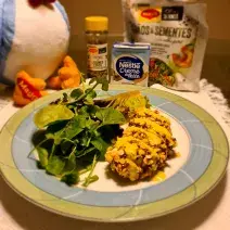 Foto aproximada de um prato redondo com bordas verde e azul com a receita de frango empanado servido com agrião. Ao fundo há produtos Maggi e Nestlé Creme de Leite e uma parte de uma pelúcia da Galinha Azul