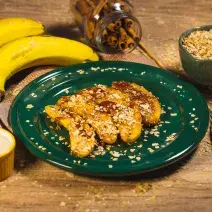 Foto da receita de banana dourada com Neston, sobre um prato verde, decorada com Neston 3 Cereais, sobre uma bancada de madeira decorada com bananas, trigo e alguns potes com açúcar e cereal dentro