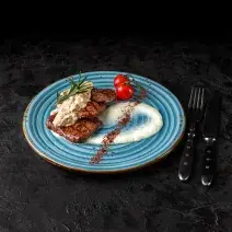 Fotografia em tons de preto em uma bancada de madeira preta. Ao centro, um prato redondo e azul contendo os medalhões e ao lado há um par de talheres.