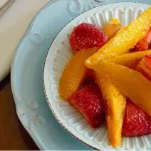 fotografia em tons de marrom, azul, amarelo e vermelho de uma bancada marrom vista de cima, contém um prato azul redondo com um pratinho redondo branco e dentro pedaços de frutas.