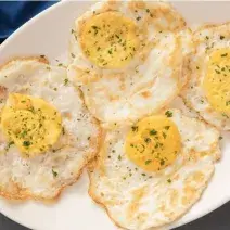 Fotografia em tons de amarelo em uma bancada de madeira escura, com um pano azul ao lado, um prato oval branco com quatro ovos fritos bem cozidos.