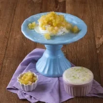 Foto da receita de pavlova de limão com abacaxi, servida em um prato azul, com creme ao lado em um pote rosa e outro pote branco com pedaços de abacaxi, tudo em uma bancada de madeira com um pano em tom violeta