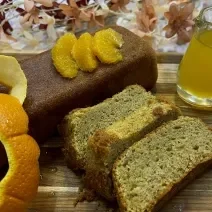 Foto da receita de Bolo de Laranja com Amêndoas. Observa-se um bolo inglês com três pedaços cortados. Como decoração, suco de laranja e a casca cortada.