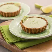 Foto da receita de tortinha de limão. Observa-se dois pratos verdes de sobremesa com as tortinhas sobre um guardanapo verde e uma tábua de madeira. Atrás, duas fatias de limão.