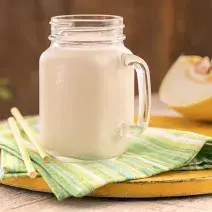 Foto em tons de amarelo da receita de smoothie de melão servida em uma jarra de vidro pequena sobre um pano listrado verde e amarelo com dois canudos ao lado. Ao fundo uma fatia grande de melão