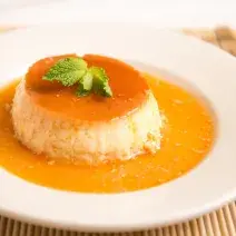 Fotografia em tons de laranja em uma bancada de madeira de cor marrom. Ao centro, um prato redondo branco contendo o gelado. Ao lado, um pano marrom claro enrolado.