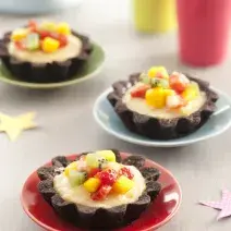 Fotografia em tons de vermelho em uma bancada de madeira clara com três pratinhos coloridos, vermelho, azul e verde e a tortinha de chocolate com frutas em cima dos pratinhos. Ao fundo, um copo rosa e um copo amarelo.