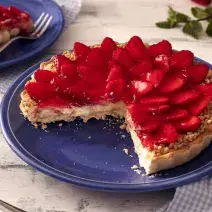 Fotografia em tons de vermelho em uma bancada de madeira de cor branca. Ao centro, um prato azul contendo a torta. Ao lado, há um pano azul quadriculado e ao fundo, um pires contendo uma fatia da torta.