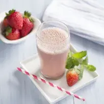 Fotografia em tons de branco e rosa tirada de cima, contém uma bancada branca com um copo transparente que contém a bebida de morango, e ao lado um morango e um canudo em tons de branco e rosa