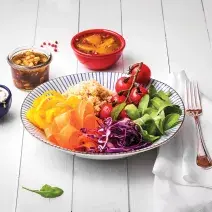 Fotografia em tons coloridos em uma bancada de madeira branca com um prato fundo com listras azuis e salada de rúcula, cenoura, cebola roxa e tomate dentro dele. Ao lado, potinhos com opções de molhos.