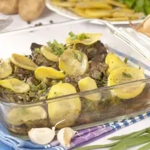 Fotografia de uma travessa de vidro quadrada com iscas de fígado e batatas, sobre um pano de mesa dobrado na cor azul com listras brancas. Ao fundo, cebolas e batatas inteiras, ao lado de um prato com legumes cortados.