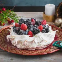 Foto da receita de pavlova natalina, branca, sobre um prato vermelho, decorada com frutas vermelhas e açúcar. Ela está numa mesa de madeira preenchida de decorações de natal como pinhas, bolas de natal e uma vela acesa ao fundo