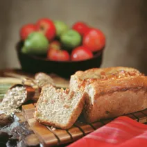 Fotografia em tons de vermelho em uma bancada de madeira com uma tábua e o pão de aveia e maçã em cima dela, com uma fatia já cortada. Ao lado, um pano vermelho e ao fundo, uma cesta com maçãs vermelhas e verdes.