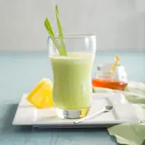 Fotografia em tons de verde em uma mesa de madeira azul clara com um prato retangular branco ao centro com um copo de vidro em cima e o suco de abacaxi, capim-santo e mel dentro dele. Ao fundo, um potinho de vidro com mel.