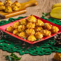 Foto da receita Batatas Natalinas, em tom amarelo e dourado, servidas em um prato vermelho quadrado sobre uma toalha verde natalina. Tudo está em uma mesa de madeira decorada com batatas cruas, pinhas natalinas e mais algumas batatas prontas.