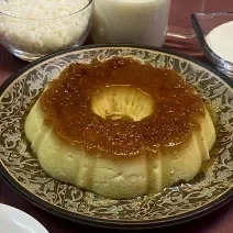Foto em tons de bege escuro da receita de pudim de arroz inteira servida sobre um prato grande decorado. Ao fundo há alguns recipientes com arroz, açúcar, leite ninho e leite MOÇA.