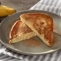 Foto em tons de cinza da receita de banana toast servida em duas metades sobre um prato de cerâmica cinza com canela em pó salpicada. Ao fundo há algumas bananas e um paninho listrado cinza