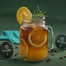 Fotografia em tons de verde de uma bancada verde, sobre ela um copo de vidro com cold brew com laranjas. Ao lado capsulas de Dolce Gusto Cold Brew e um paninho azul.